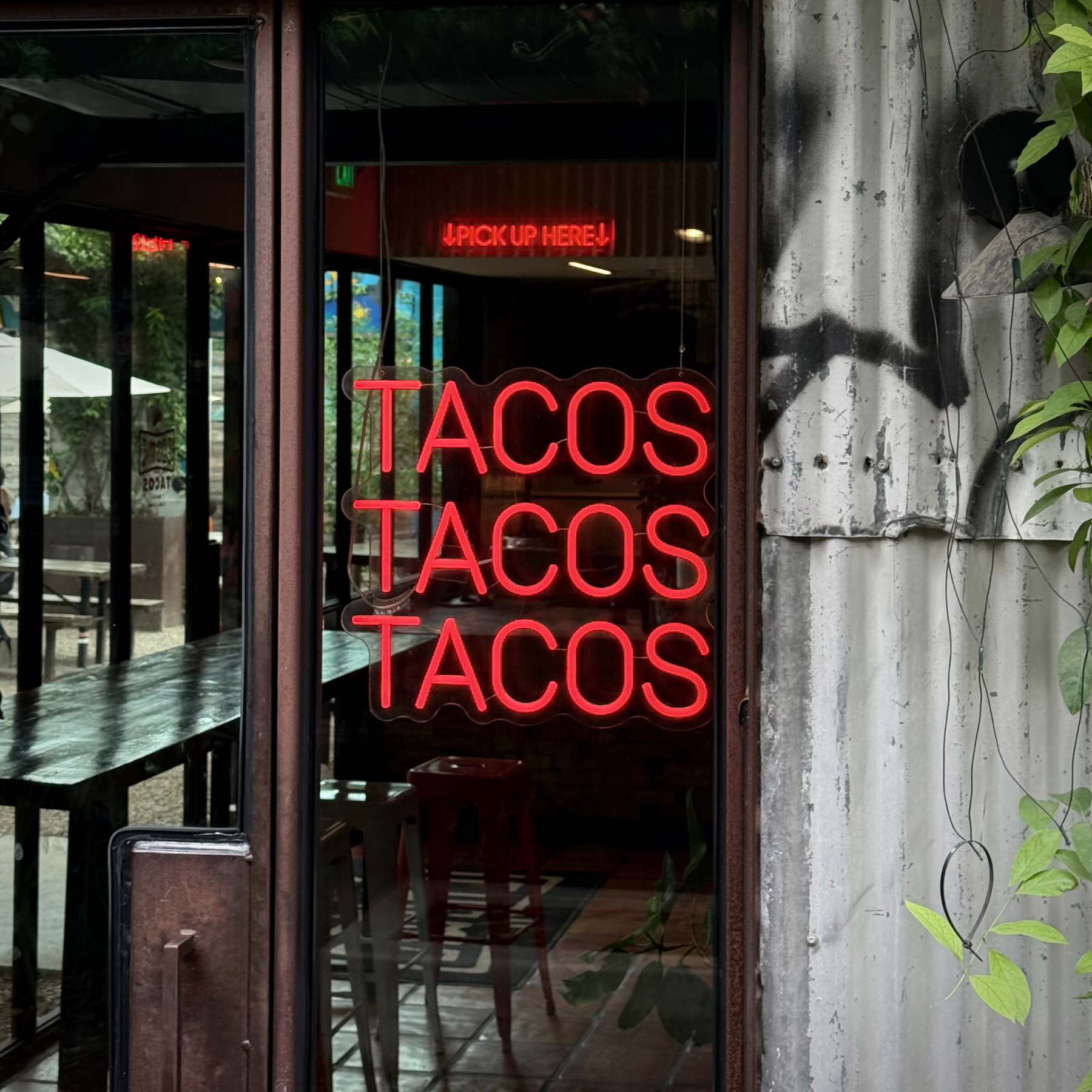 A neon sign displaying the word TACOS three times is mounted on a window next to a rustic metal wall.