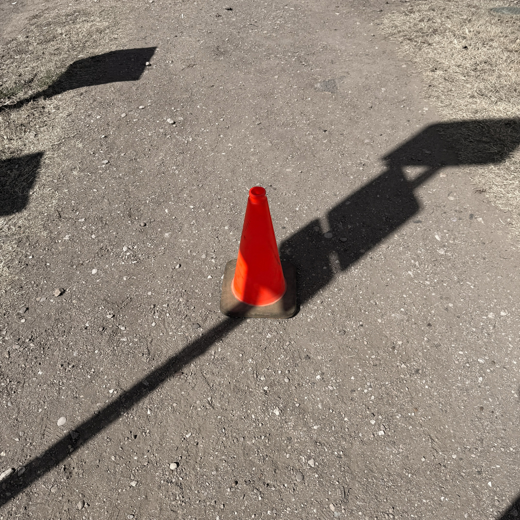 A single bright orange traffic cone is placed on a rough, dusty surface with shadows cast to the side.