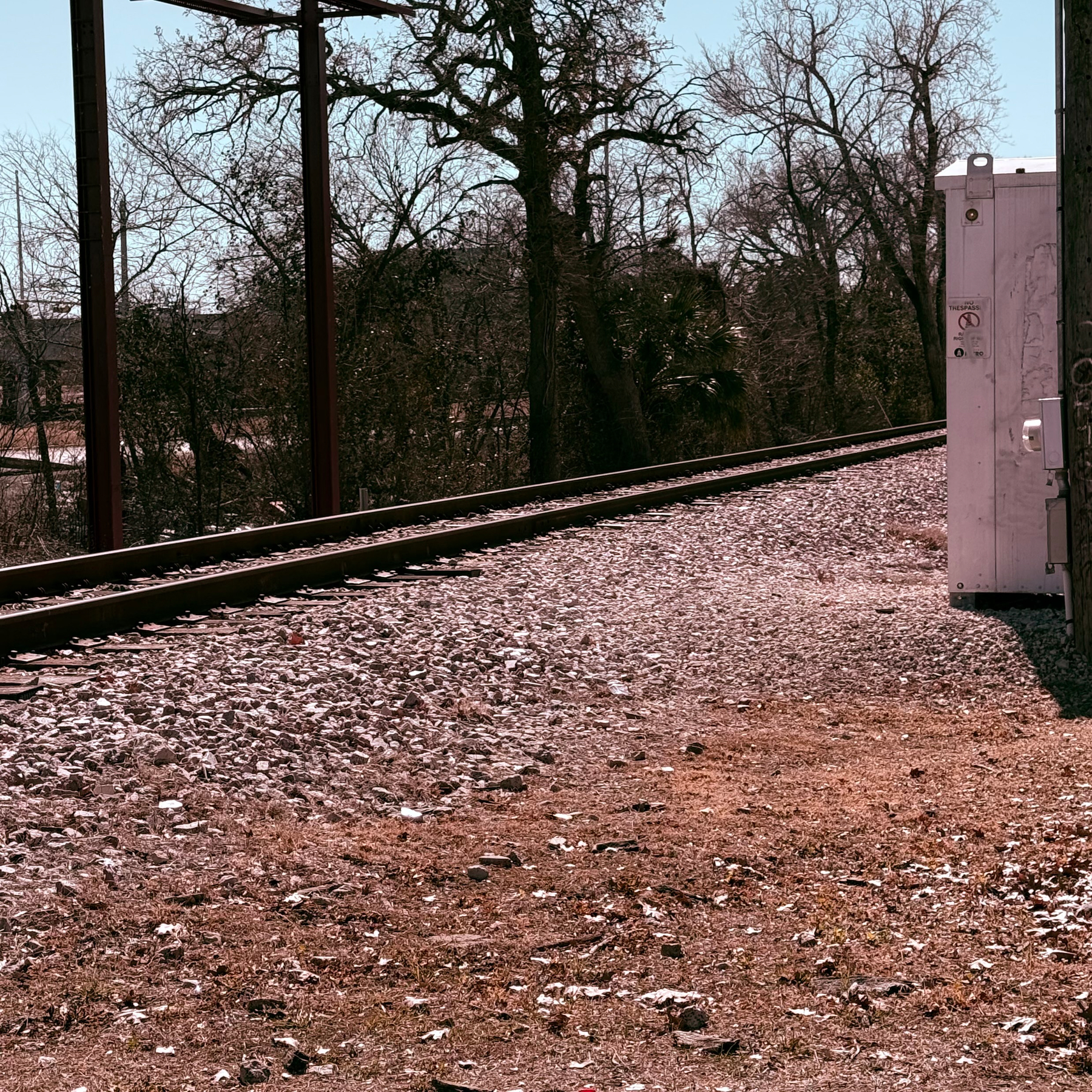 A gravelly train track runs parallel to a grassy area with a utility box, surrounded by bare trees and clear skies.